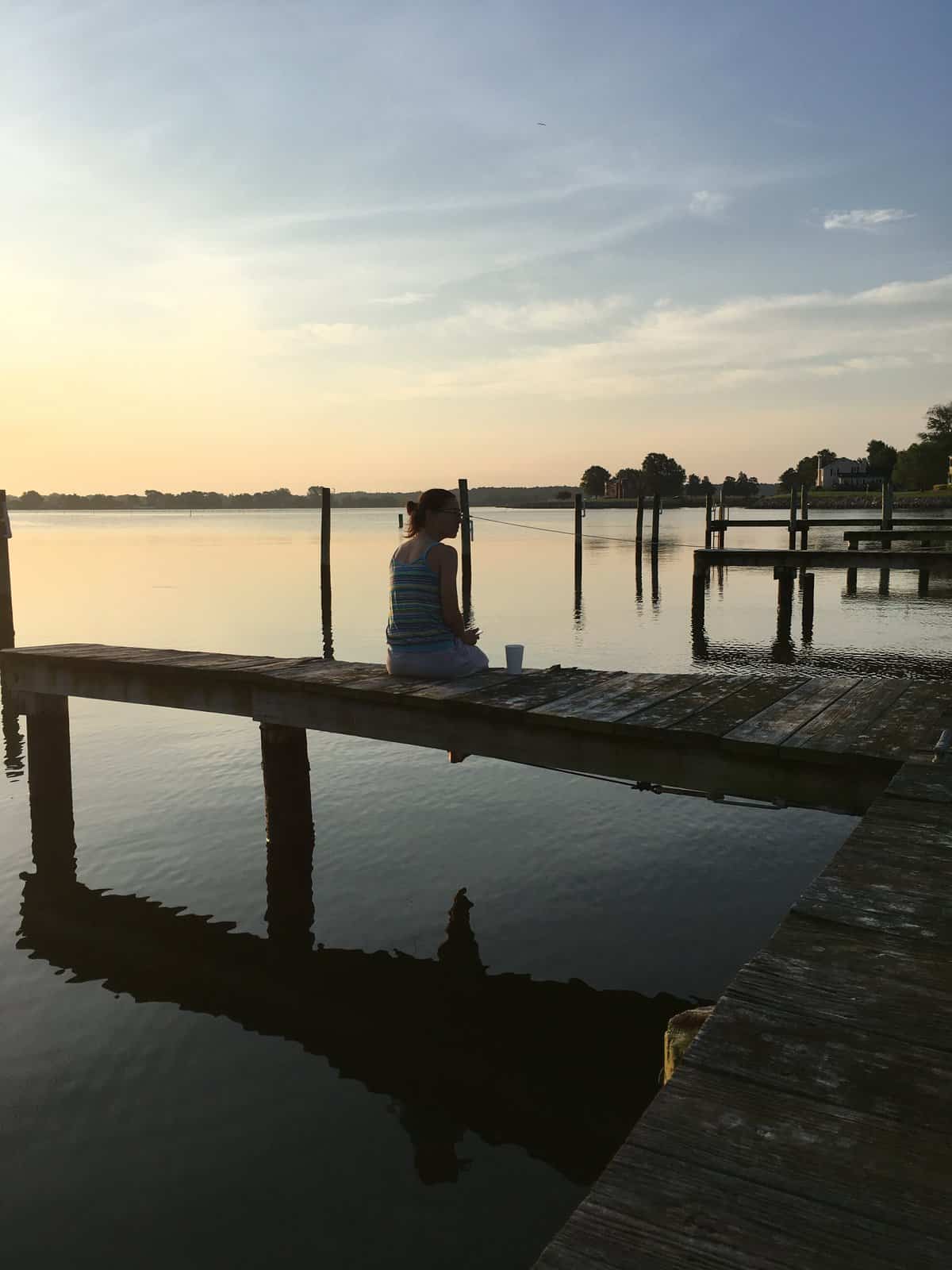 a woman sitting on the docks with a cup of coffee, at sunrise