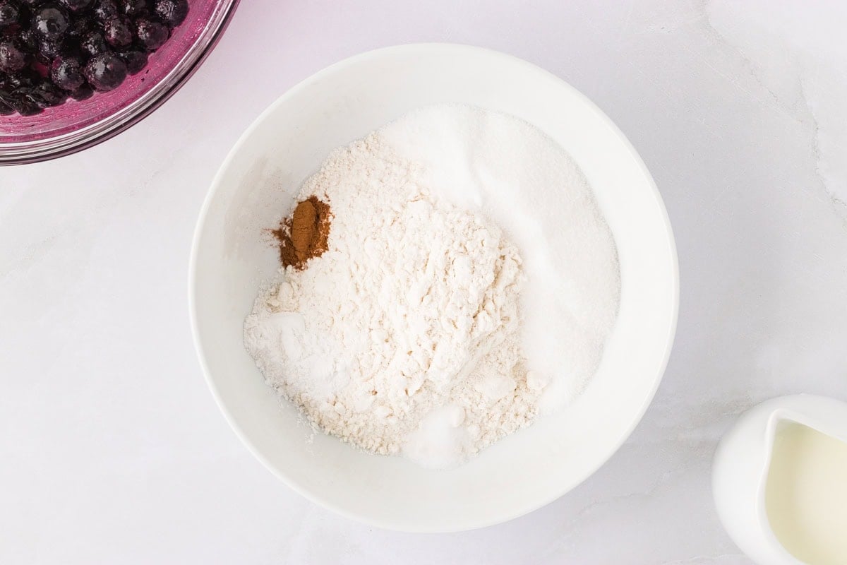 Gluten-free flour, sugar, baking powder, cinnamon, and salt mixed in a white bowl with a pitcher of milk nearby.