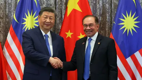 Getty Images China's President Xi Jinping and Malaysia's Prime Minister Anwar Ibrahim smile and shake hands at the official residence of the prime minister in Putrajaya. They both wearing suits and blue ties.