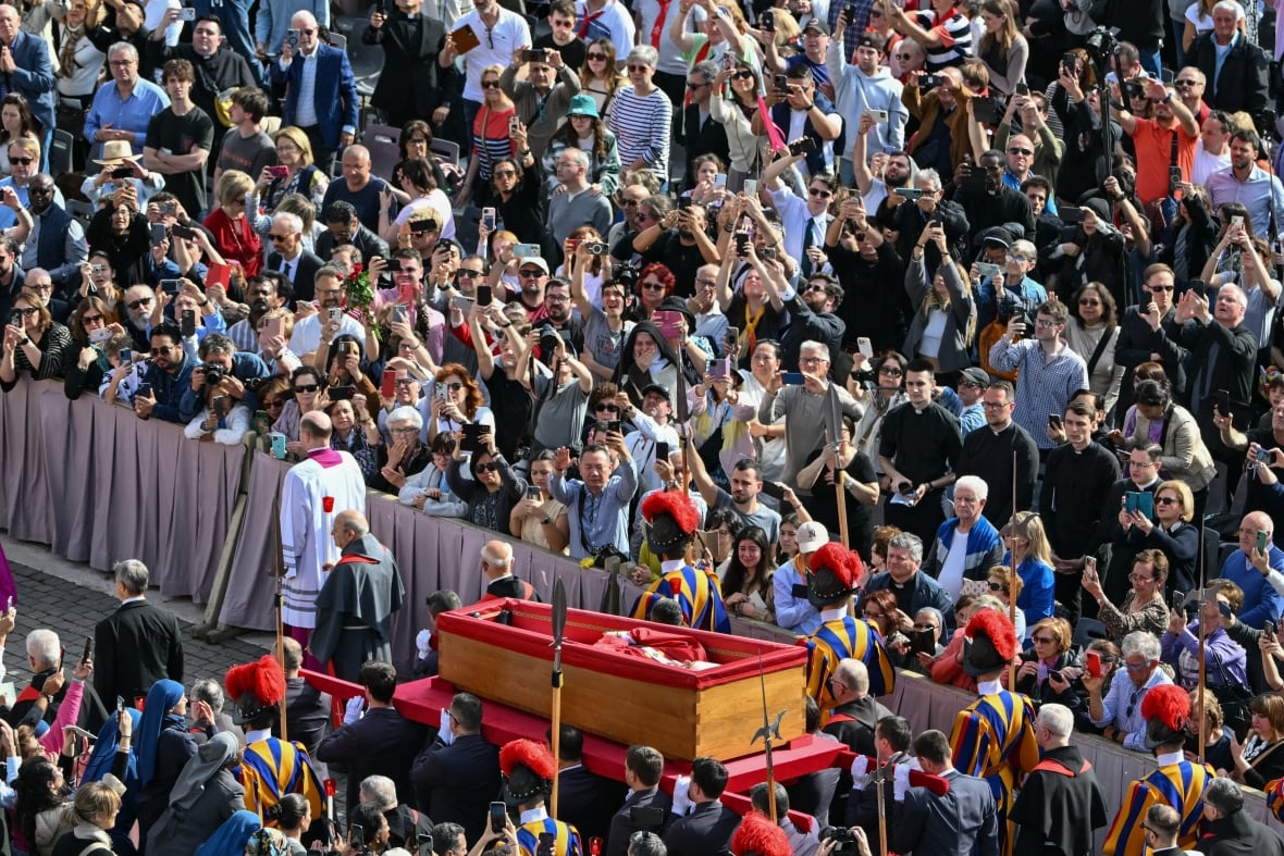 Throngs of people are shown lineup up behind barriers as a procession with a casket passes by.