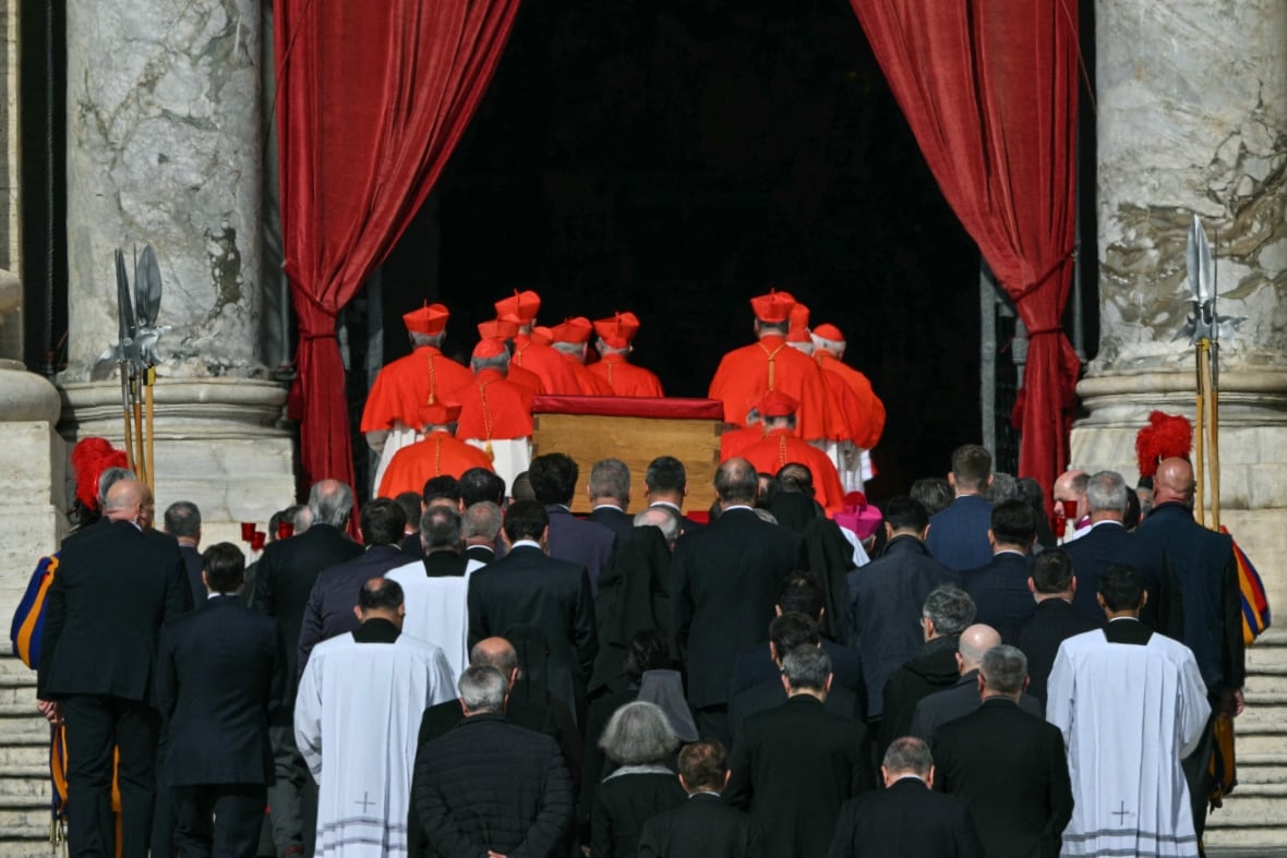 Dozens of figures in religious garments are shown from behind near a casket, as the large doors to a church are opened.