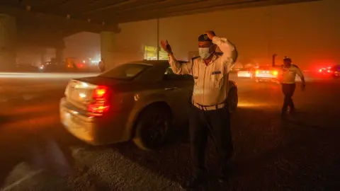 Getty Images Cars with red headlights drive through the orange sandstorm on a busy road in Najaf, Iraq on 14 April 2025. Police wearing face masks and white shirts gesture to direct traffic in the orange cloud of the sandstorm.