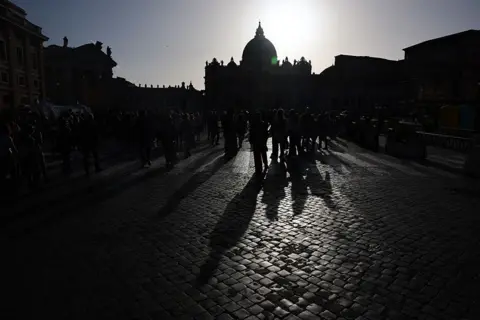 NurPhoto via Getty Images A silhouetted image of people standing in St Peter's square at the Vatican after the Pope's death.
