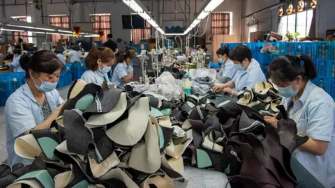 Getty Images Female workers in blue face masks and blue uniforms, seated at a long table in a shoe factory in Hunan, China. They are working on a pile of brown boots.