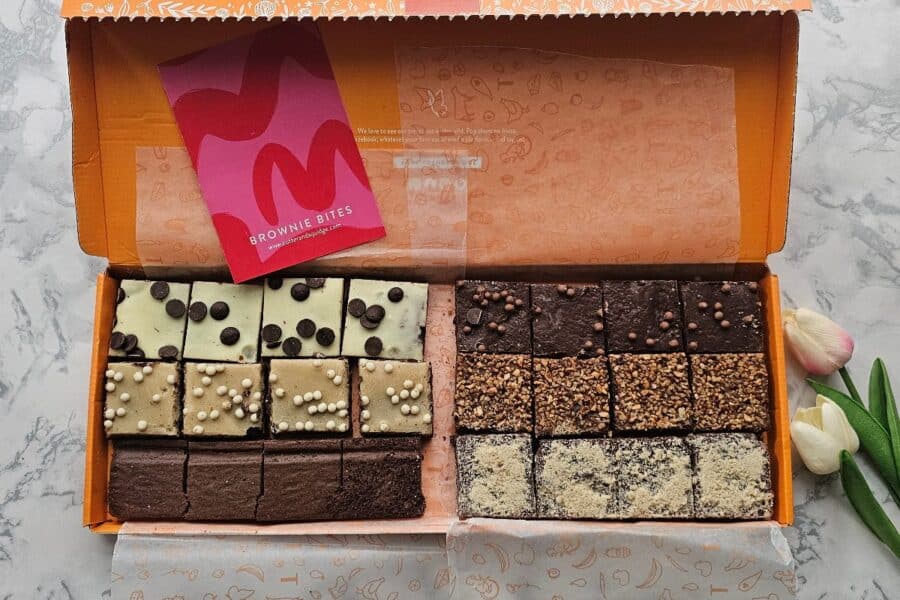 An open box of Brownie Bites from Cutter & Squidge, showcasing an assortment of delicious brownies in various flavours, neatly arranged in rows. A bright pink "Brownie Bites" card rests on top, with artificial tulips beside the box on a marble surface.