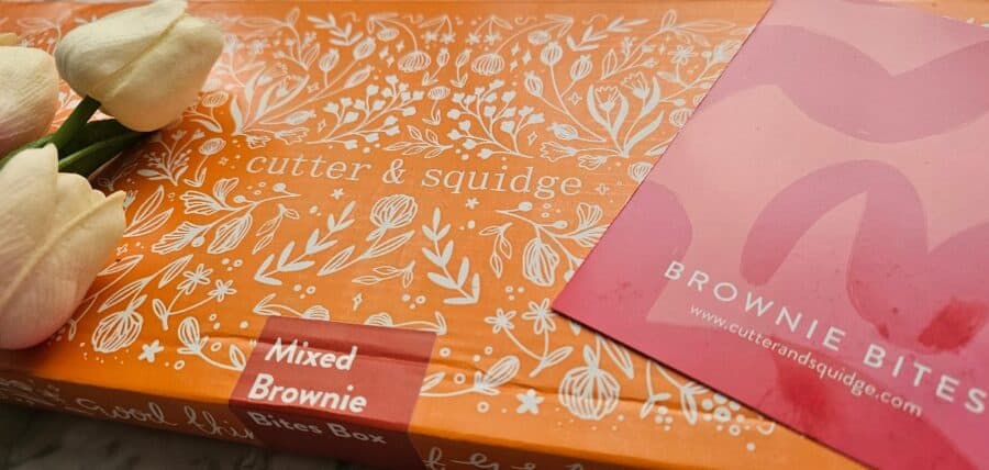 A beautifully designed Cutter & Squidge Mixed Brownie Bites Box with an intricate floral pattern on its orange packaging, accompanied by a pink "Brownie Bites" card and artificial tulips.