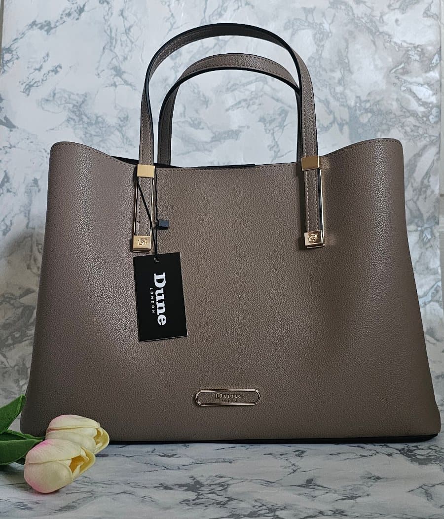 A stylish Dune London Dorry Tote Bag in Neutral with gold hardware, displayed upright on a marble surface with pink and cream tulips—perfect gift inspiration for Mother’s Day.