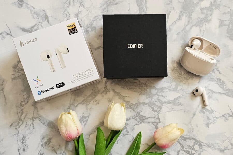 A set of ivory coloured Edifier W320TN wireless earbuds displayed with their packaging, including a white retail box and a sleek black gift box, alongside artificial tulips on a marble surface.