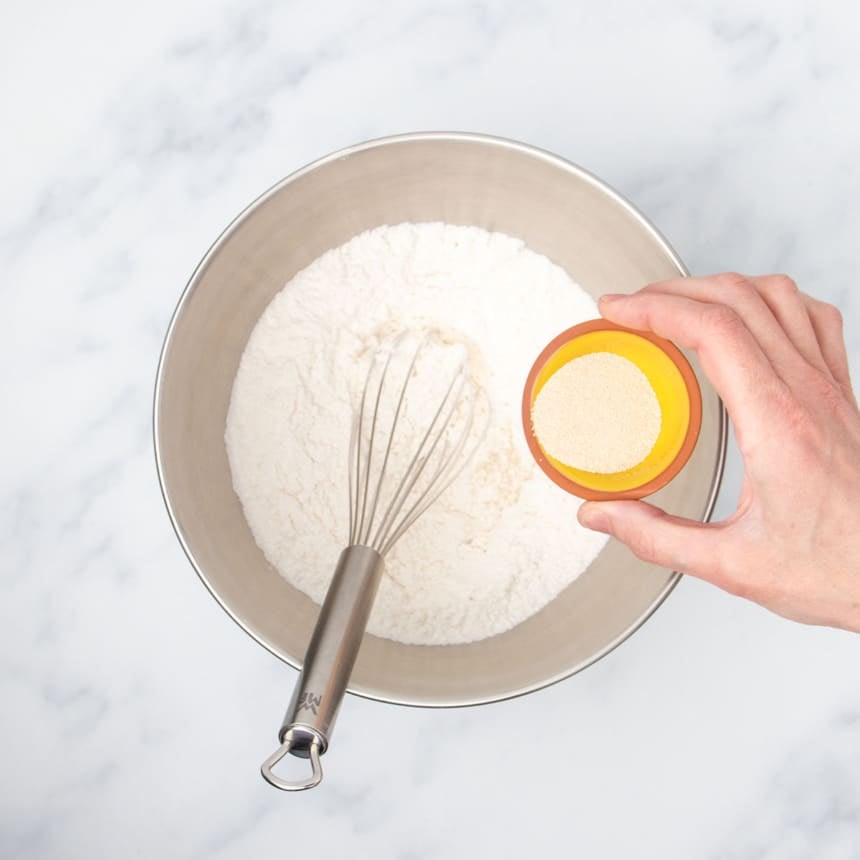 large metal mixing bowl with white flour inside with metal wire whisk and hand pouring yeast from small yellow bowl