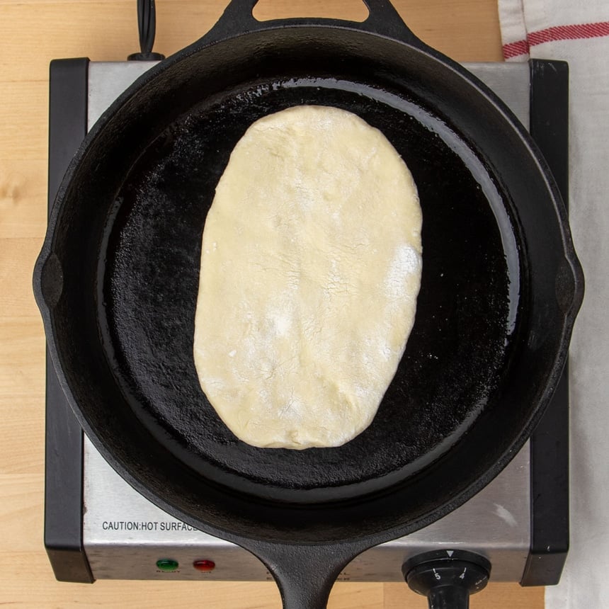 black round cast iron skillet with one raw oval of dough cooking