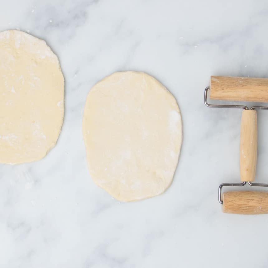 two floured flat oval pieces of raw dough with small blond wood roller