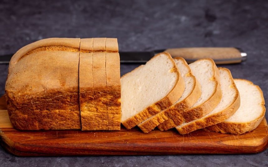 side image of loaf of bread with brown crust and white crumb with 8 slices cut, and whole thing sitting on a brown bread board, with a bread knife in background