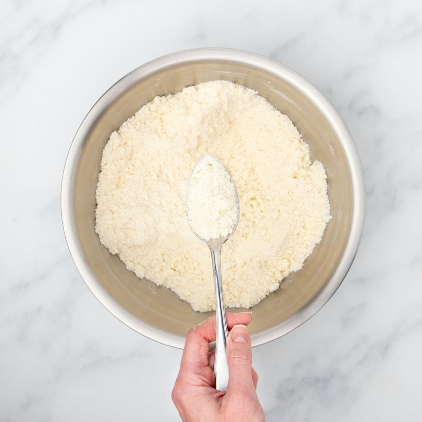 hand holding metal mixing spoon with sandy pale tan mixture of butter mixed with flour, and more of the mixture in the same mixing bowl