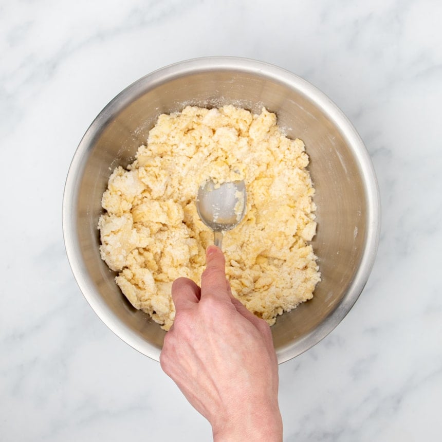 hand pressing metal mixing spoon into sandy mixture in bowl with beaten egg added, pressing mixture into clumps