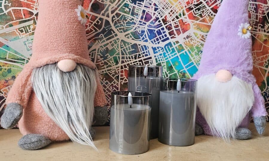A set of three grey LED votive candles from Pagazzi, placed between two pastel gnomes with daisy decorations, in front of a colourful map-themed background, ideal gift inspiration for Mother’s Day.