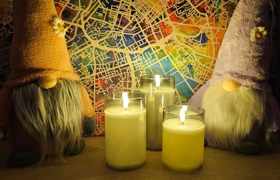 A glowing set of Pagazzi grey LED votive candles lit in a dark room, placed between two gnomes with daisy hats—perfect cosy gift inspiration for Mother’s Day.