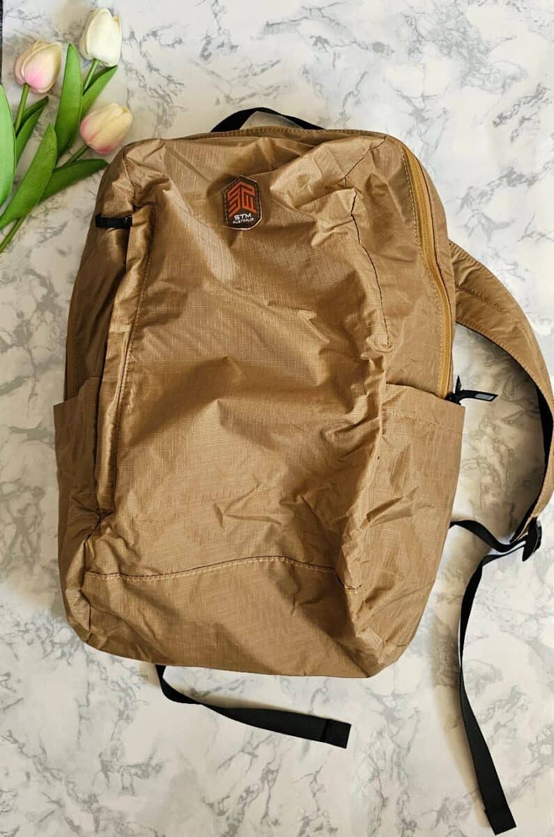 A STM Goods backpack in Coffee colour, featuring a lightweight, durable design with multiple compartments, displayed on a marble surface with artificial tulips.