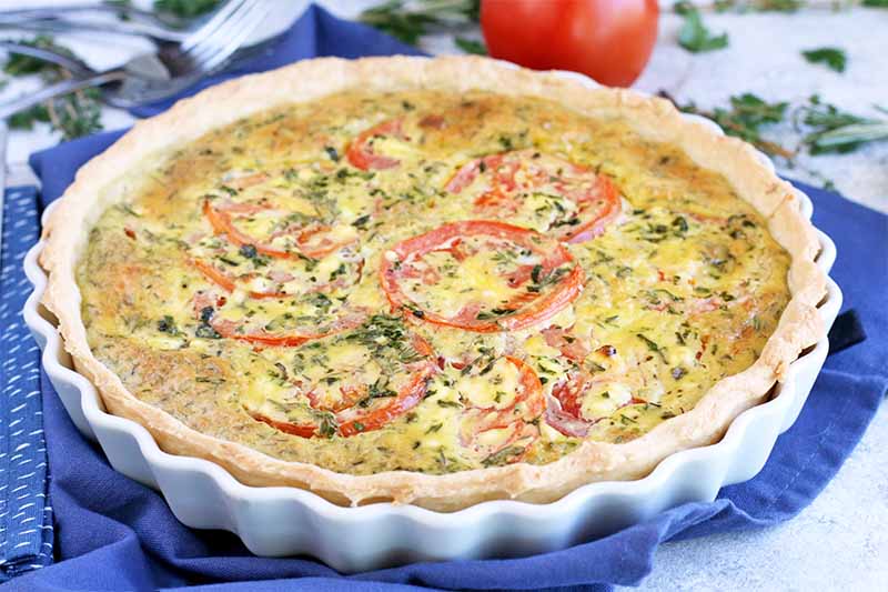A close up of a freshly baked tomato and goat cheese tart in a white ceramic dish, set on a blue fabric.