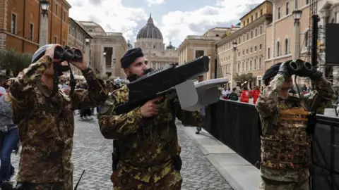 EPA Italians soldiers provide security at the Vatican. One holds holding a large black anti-drone device while another looks through binoculars.