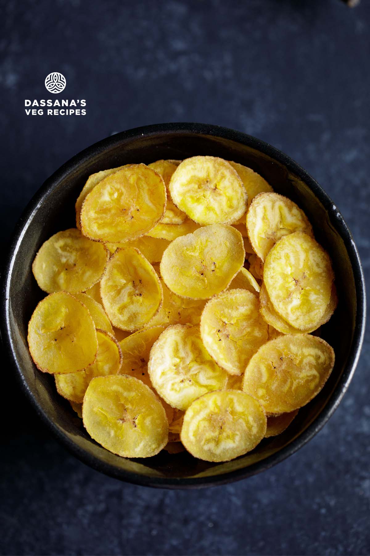 banana chips or wafers served in a black bowl on a dark bluish black wooden board.