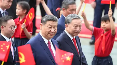 Getty Images Vietnam's General Secretary of the Communist Party To Lam (R) receives China's President Xi Jinping during a ceremonial welcome at the Presidential Palace in Hanoi on April 14, 2025. Both men are in dark suits as children around them wave Vietnam's flag.