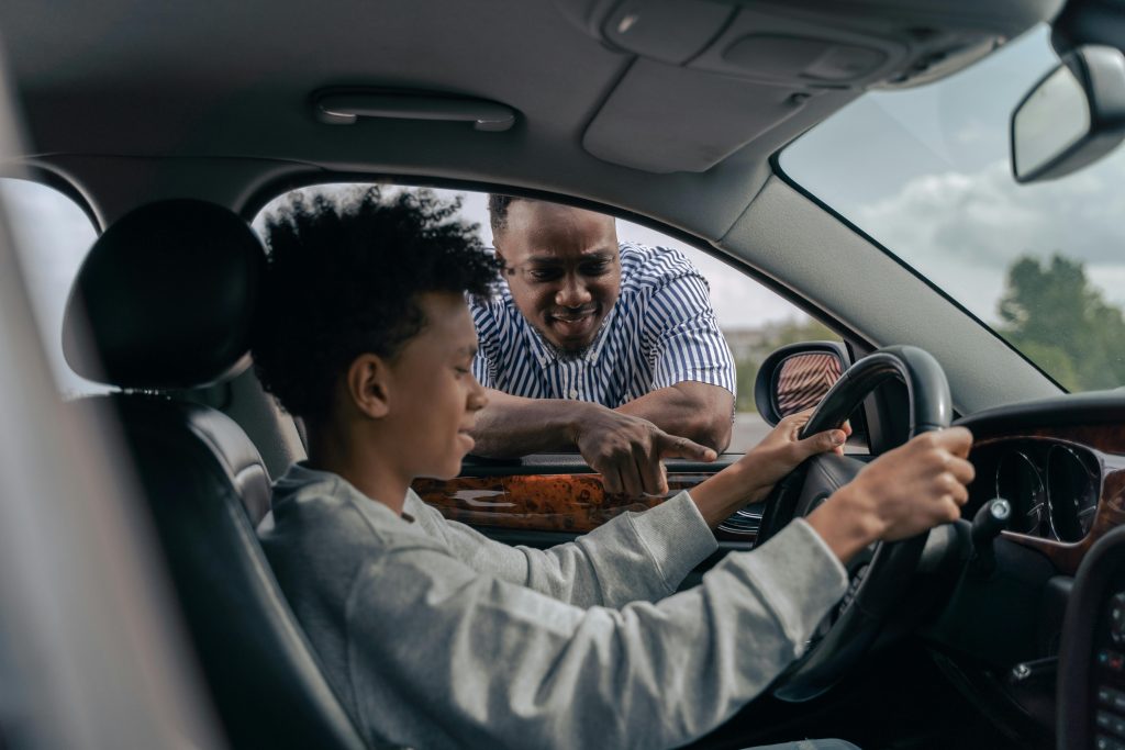 Dad looks in window as boy takes steering wheel of a car.  Photo by Ron Lach for pexels.