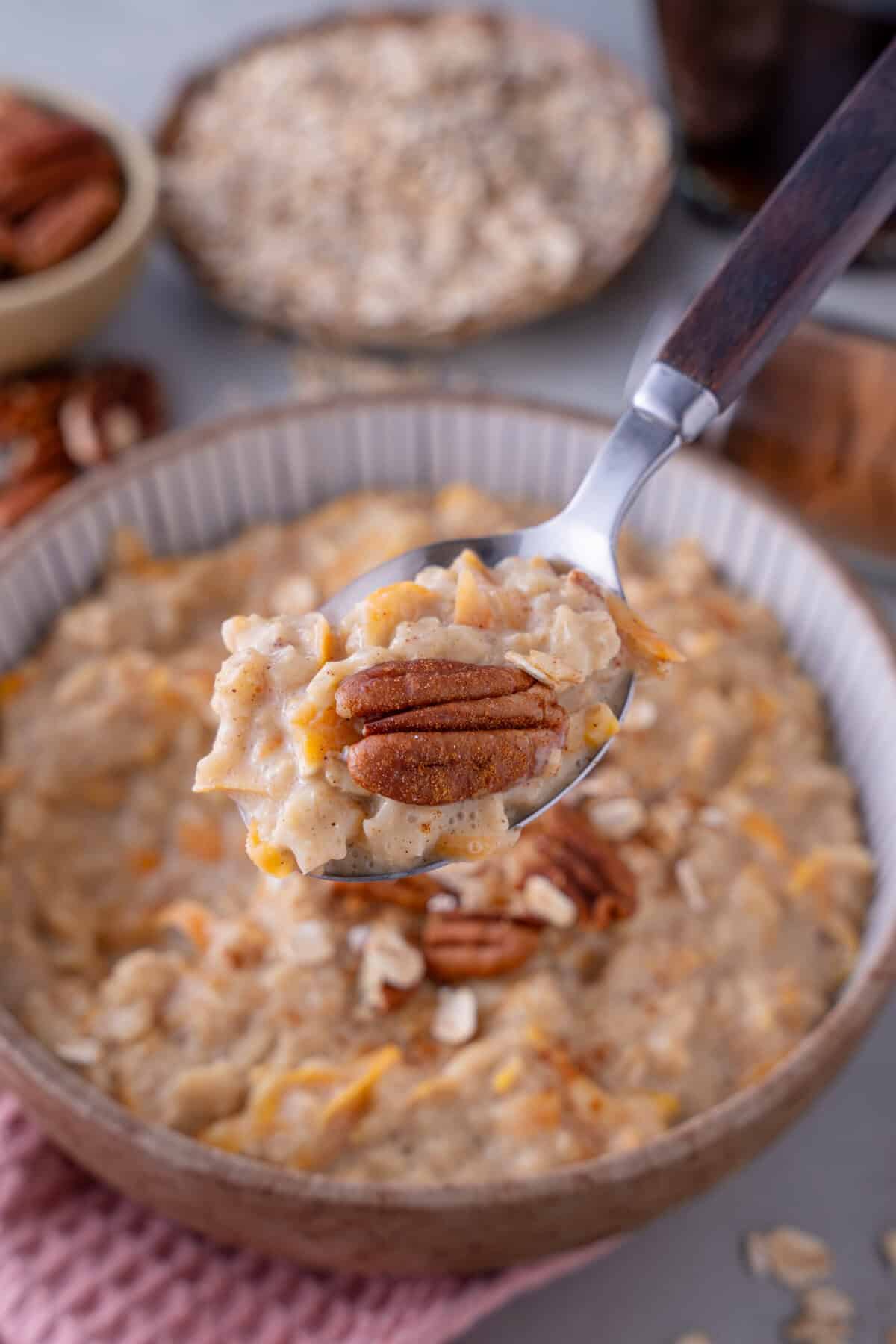 A spoonful of carrot cake porridge with a bowl of porridge behind, topped with pecan nuts.