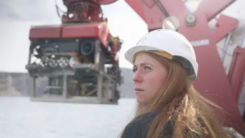 Kevin Church/BBC A photograph of Professor Isobel Yeo wearing a hard white hat on the deck of RRS Discovery with a red robot suspended in the air behind her, waiting to be lowered into water underneath.