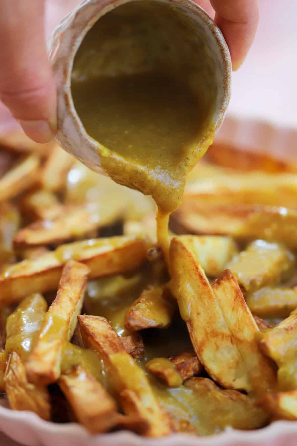 Hand pouring a jug of chip shop curry sauce over a bowl of homemade chips.