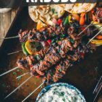 A tray with grilled lamb kabobs, pita bread, grilled vegetables, and a bowl of white sauce. Text reads: "How to make grilled lamb kabobs. Get the full recipe on girlcarnivore.com.