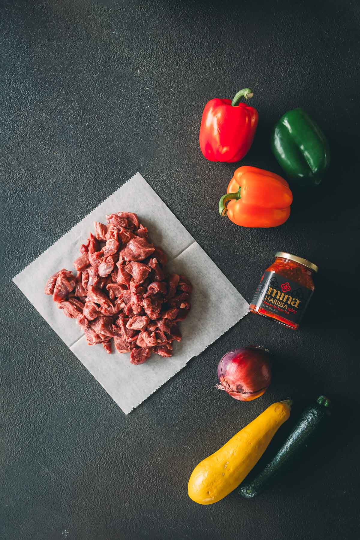 Raw subed lamb on parchment paper next to a red bell pepper, orange bell pepper, green bell pepper, red onion, yellow squash, zucchini, and a jar of harissa on a dark surface.