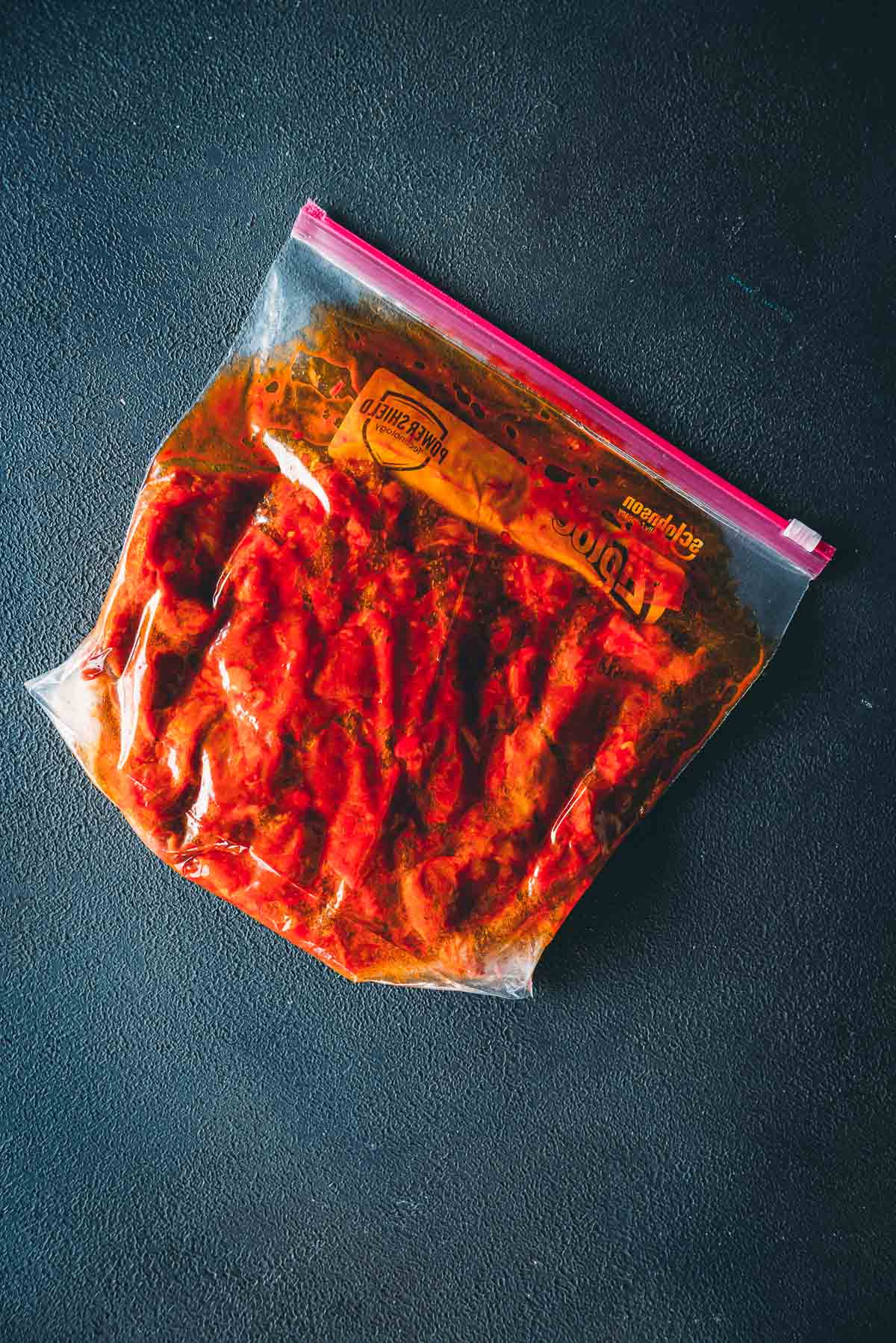 A resealable plastic bag filled with marinated lamb in a harissa, placed on a dark textured surface.