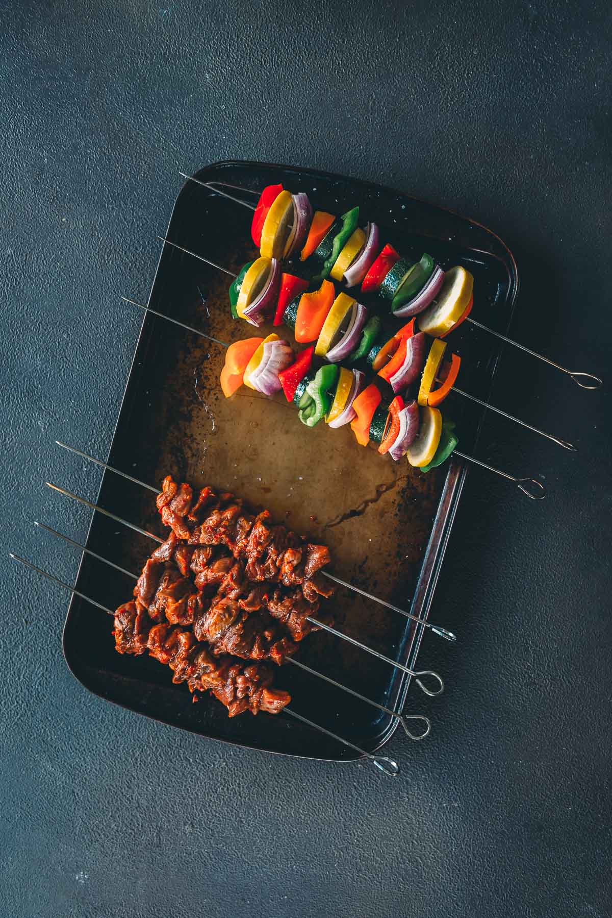 Tray with metal skewers holding marinated lamb pieces and colorful chopped vegetables including bell peppers and onions, arranged on a dark surface.