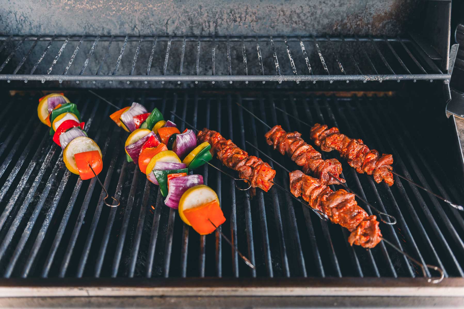Skewers on a grill with assorted vegetables and with marinated lamb, all cooking over open grates inside a barbecue grill.