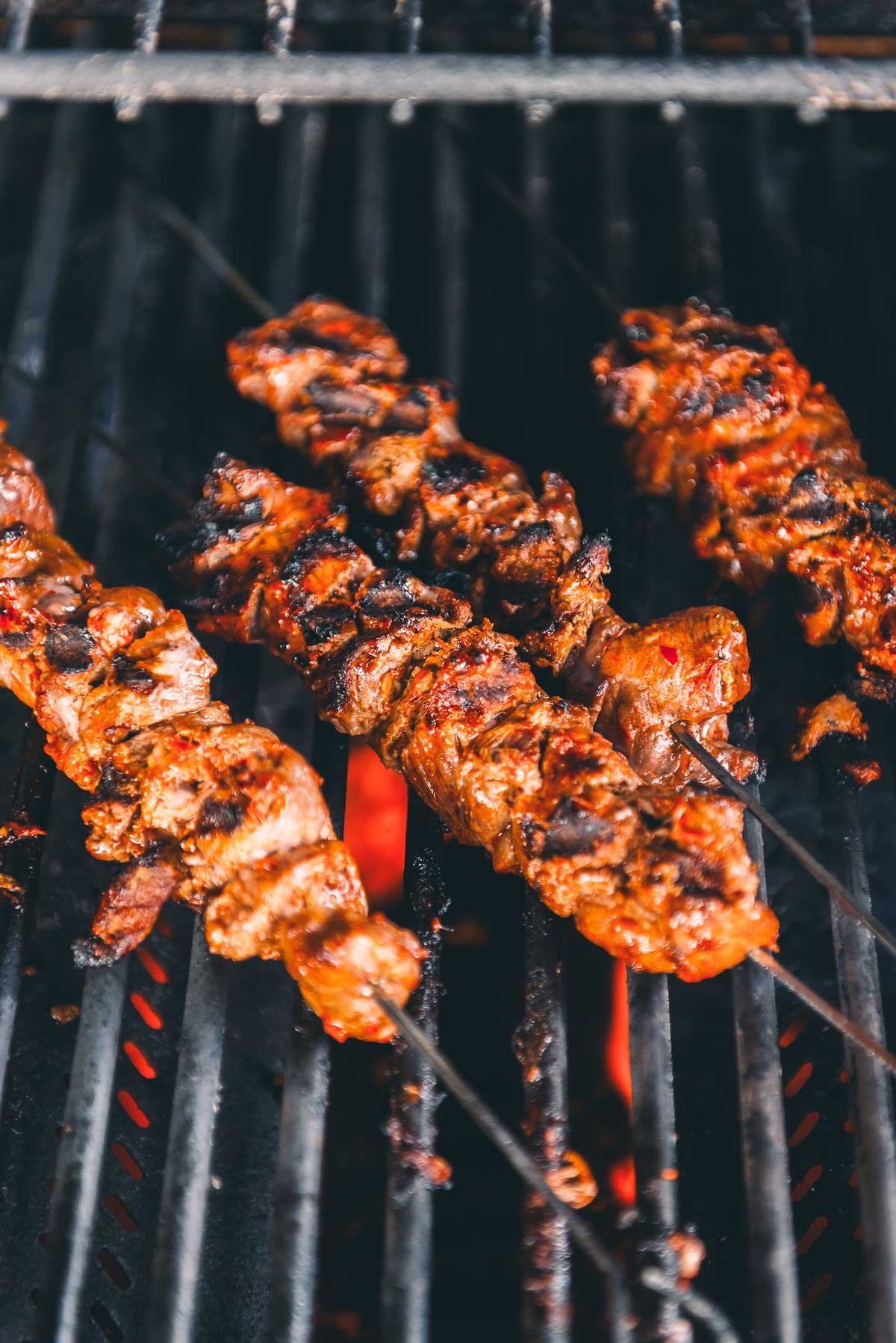 Four skewers of marinated lamb are grilling over open flames on a barbecue grill.