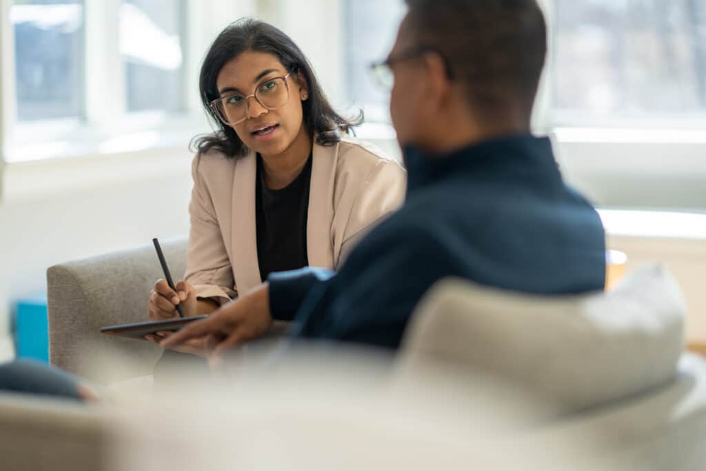 A middle aged gentleman sits across from his female therapist as they talk through his struggles.  The Therapist is dressed professionally and taking notes on a tablet.