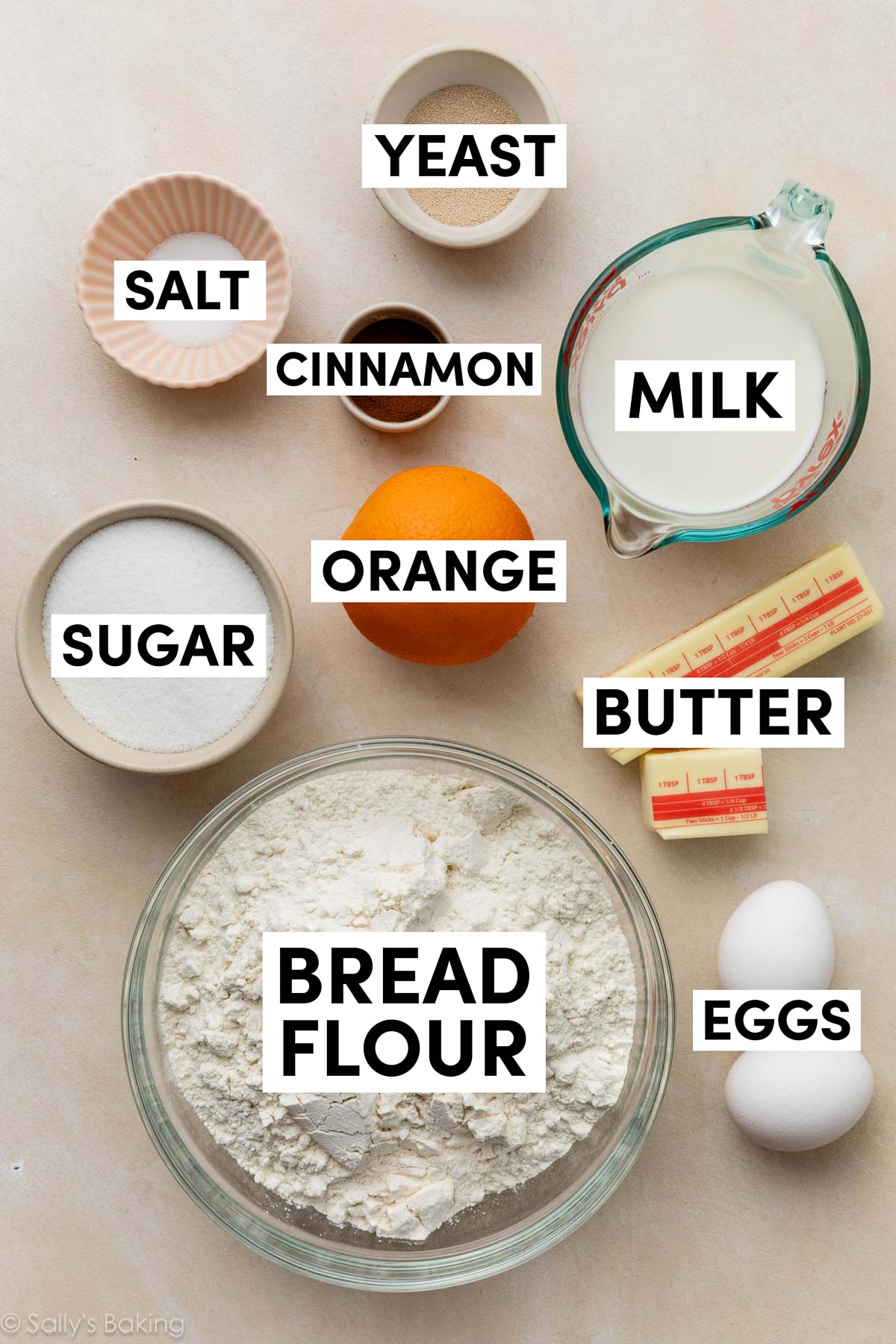 ingredients on beige surface including bread flour, butter, milk, eggs, cinnamon, sugar, and yeast.