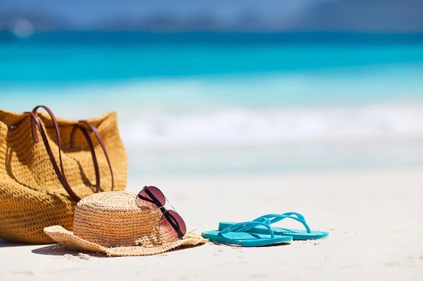 A bag, hat and flip flops in the sand at the beach with the blue water in the background.