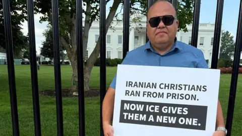 Getty Images Torosian stands in front of the White House gate holding a sign that says: Iranian Christians ran from prison. Now ICE gives them a new one.