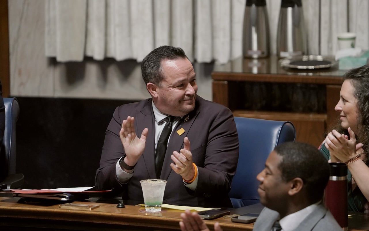 A man in a suit applauds while sitting in a legislative chamber.
