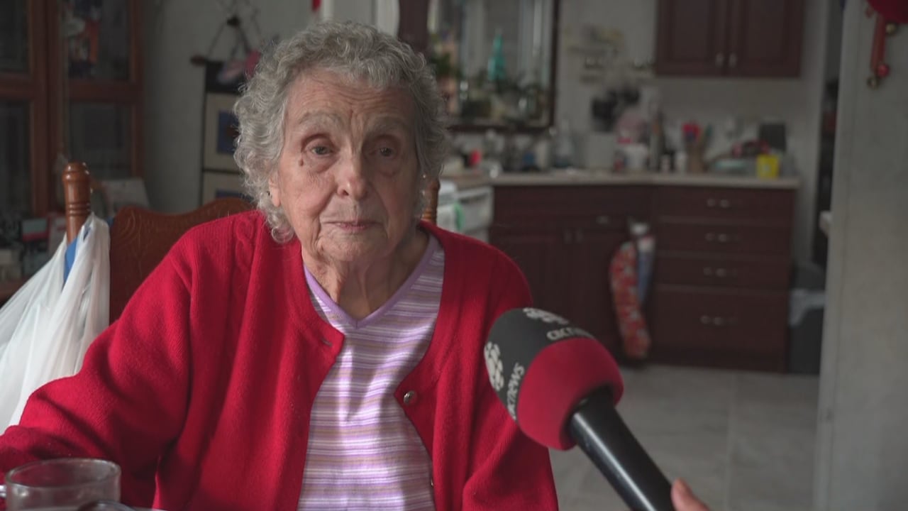 An elderly woman in a red sweater being interviewed inside her home.