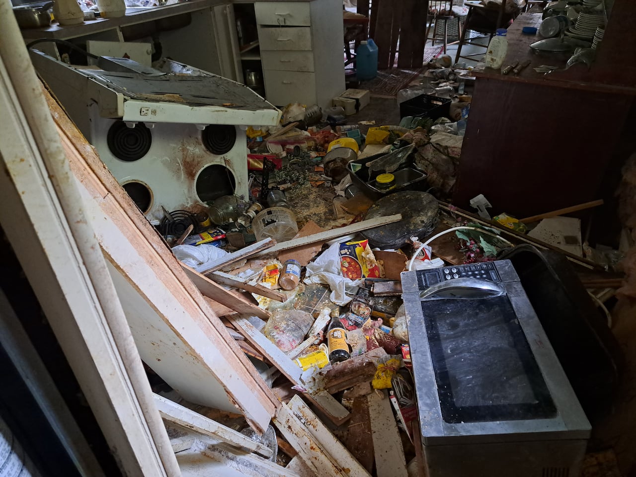 A destroyed kitchen filled with debris, broken cabinets, scattered food, and overturned appliances strewn across the floor.