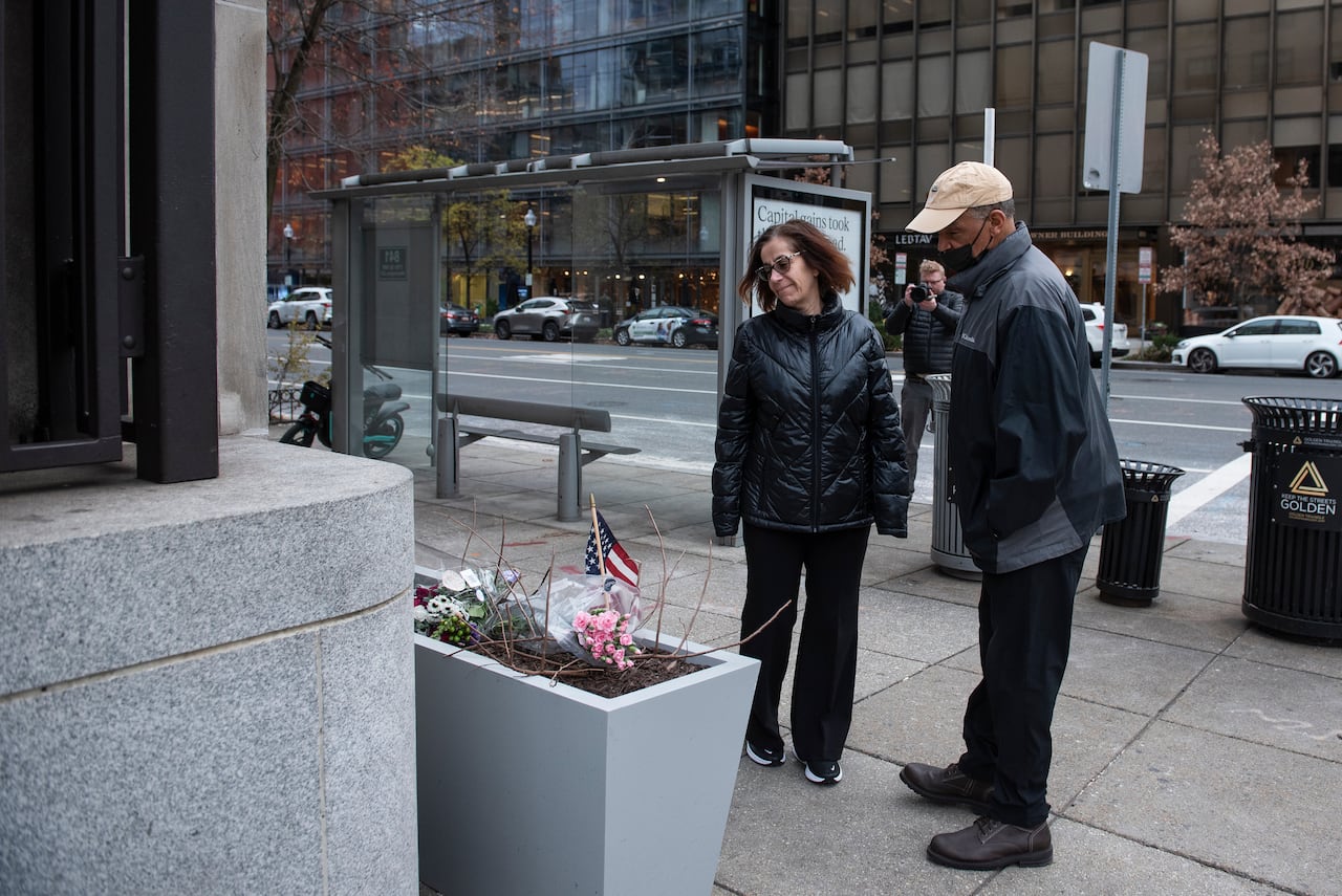 Two people, a man and a woman, look at a makeshift memorial on a sidewalk that includes a U.S. flag.