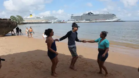 Reuters Cruise ships, used as accommodation for COP30 in the river. Three people stand on the beach in front of them.