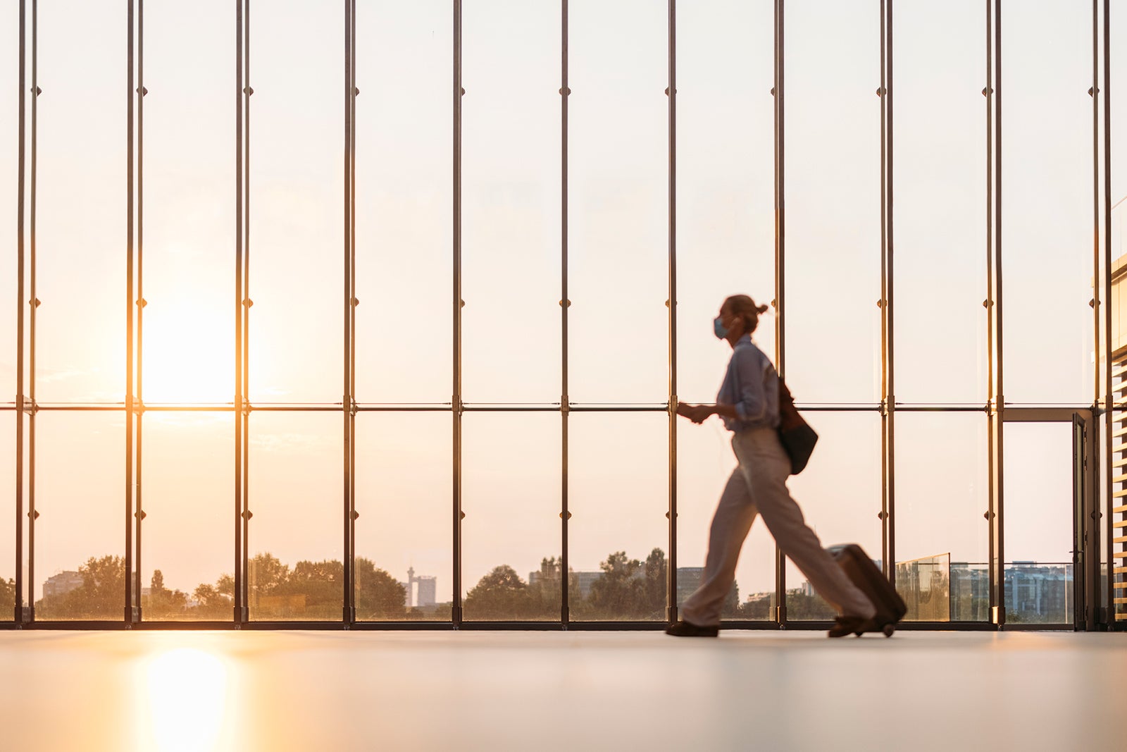 Out of Focus: Business woman with protective face mask and luggage walking at terminal hall with big window, traveling during coronavirus pandemic