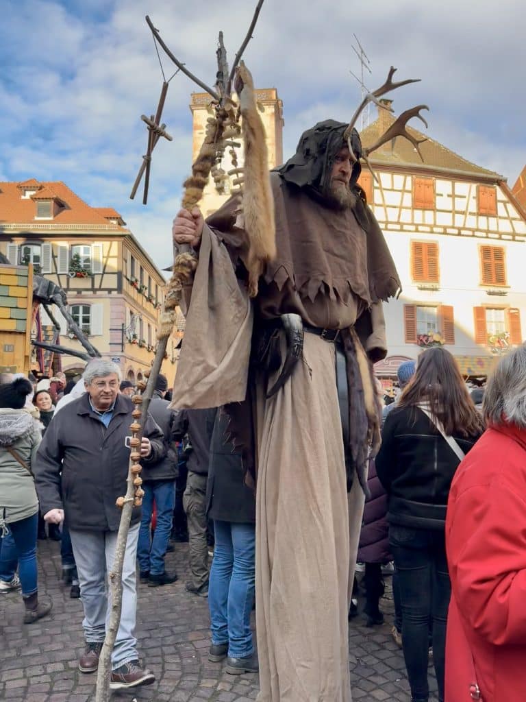 A man on stilts wearing a cloak, holding a tall cane, and wearing a hood and a headrest with antlers on it.