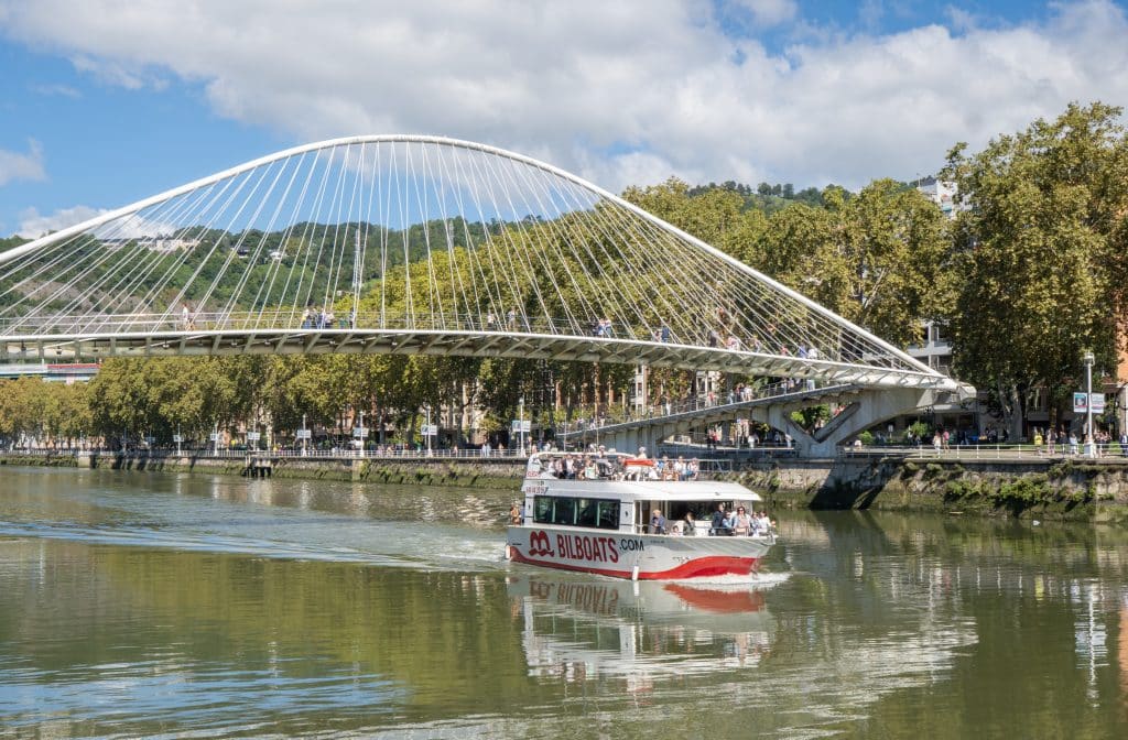 A boat filled with tourists going down the river in Bilbao, beneath the white spindly bridge.