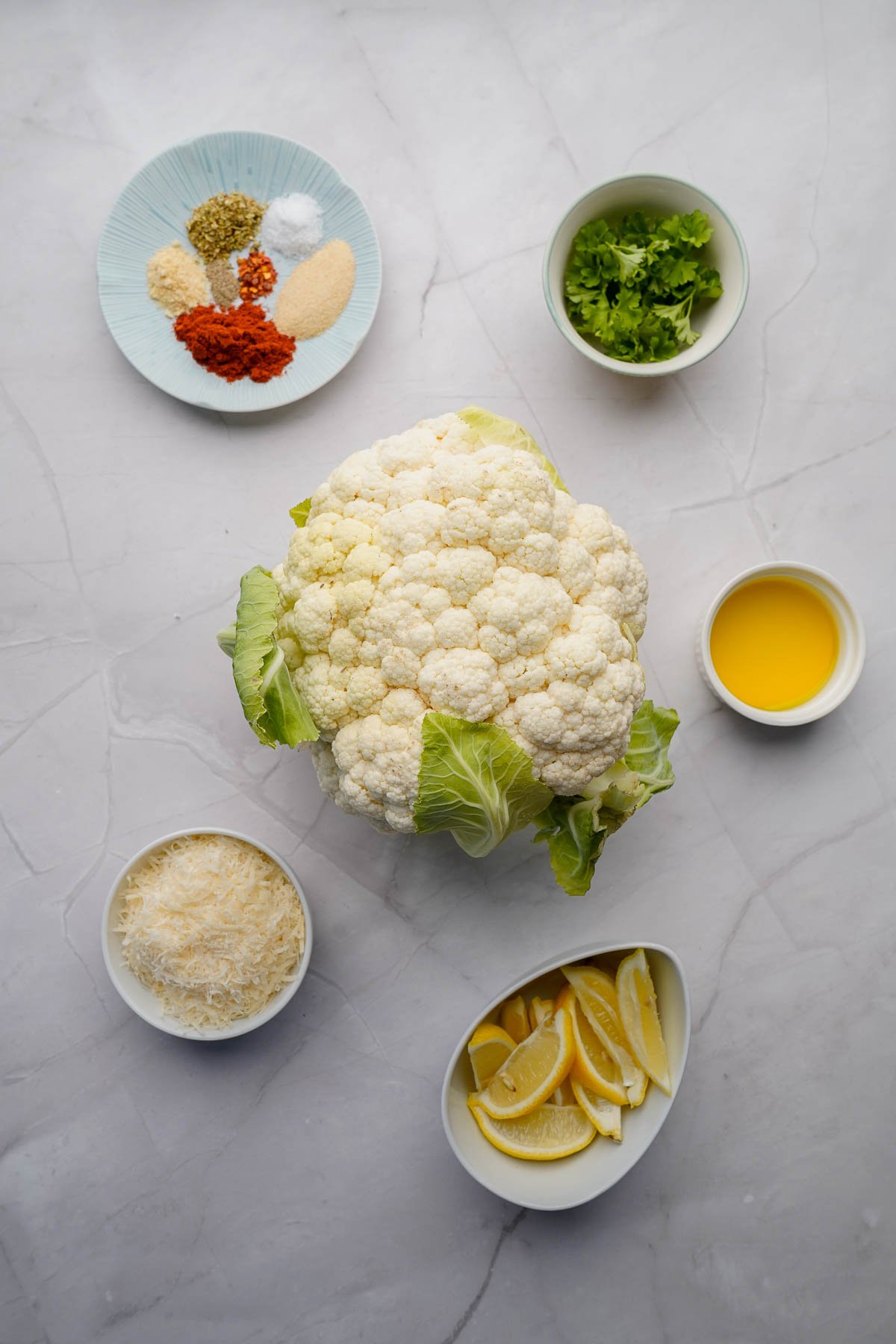 Overhead view of a whole cauliflower surrounded by small bowls containing spices, chopped herbs, grated cheese, lemon wedges, and oil on a light surface.