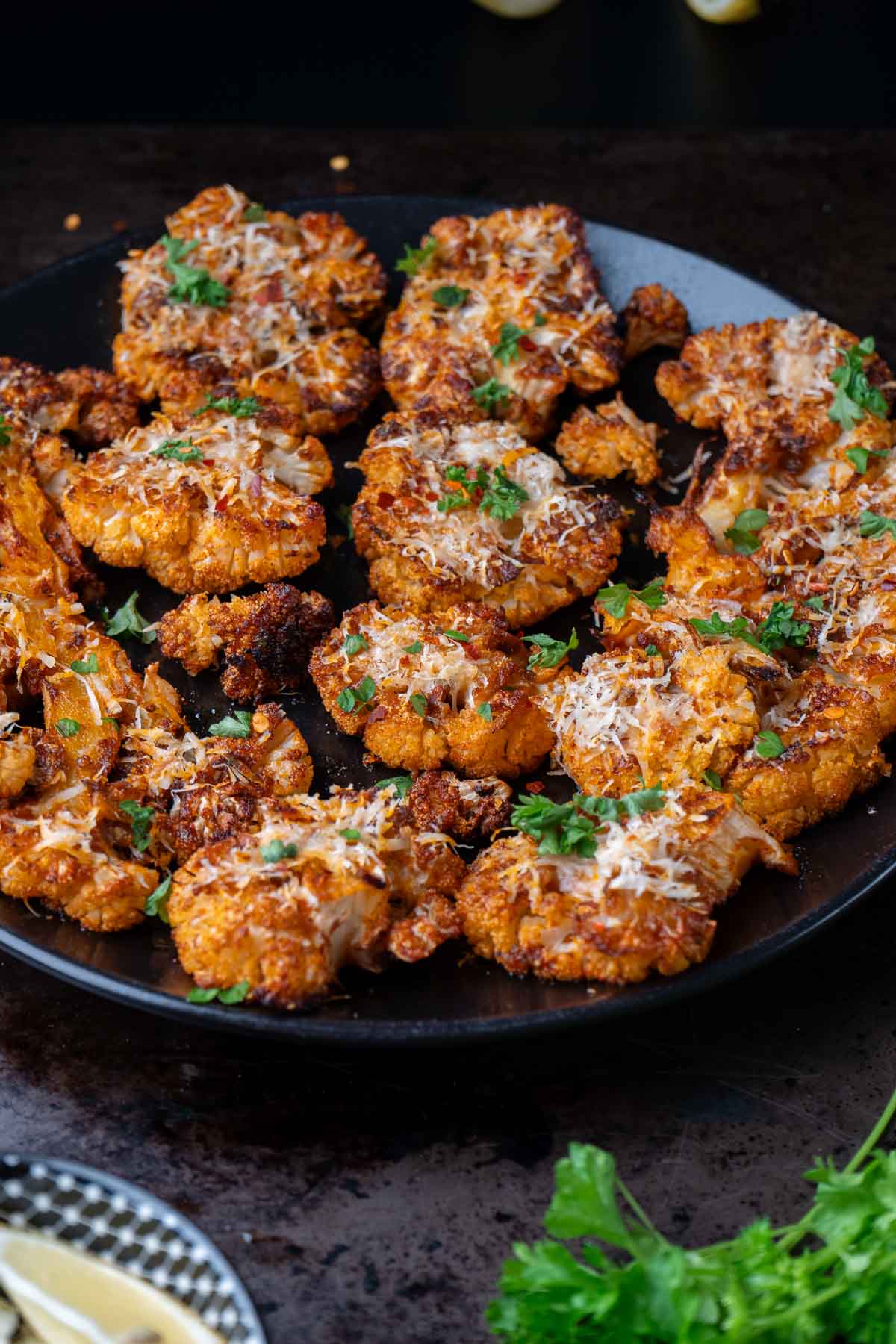 A black plate filled with roasted cauliflower steaks topped with grated cheese and chopped herbs, placed on a dark surface.