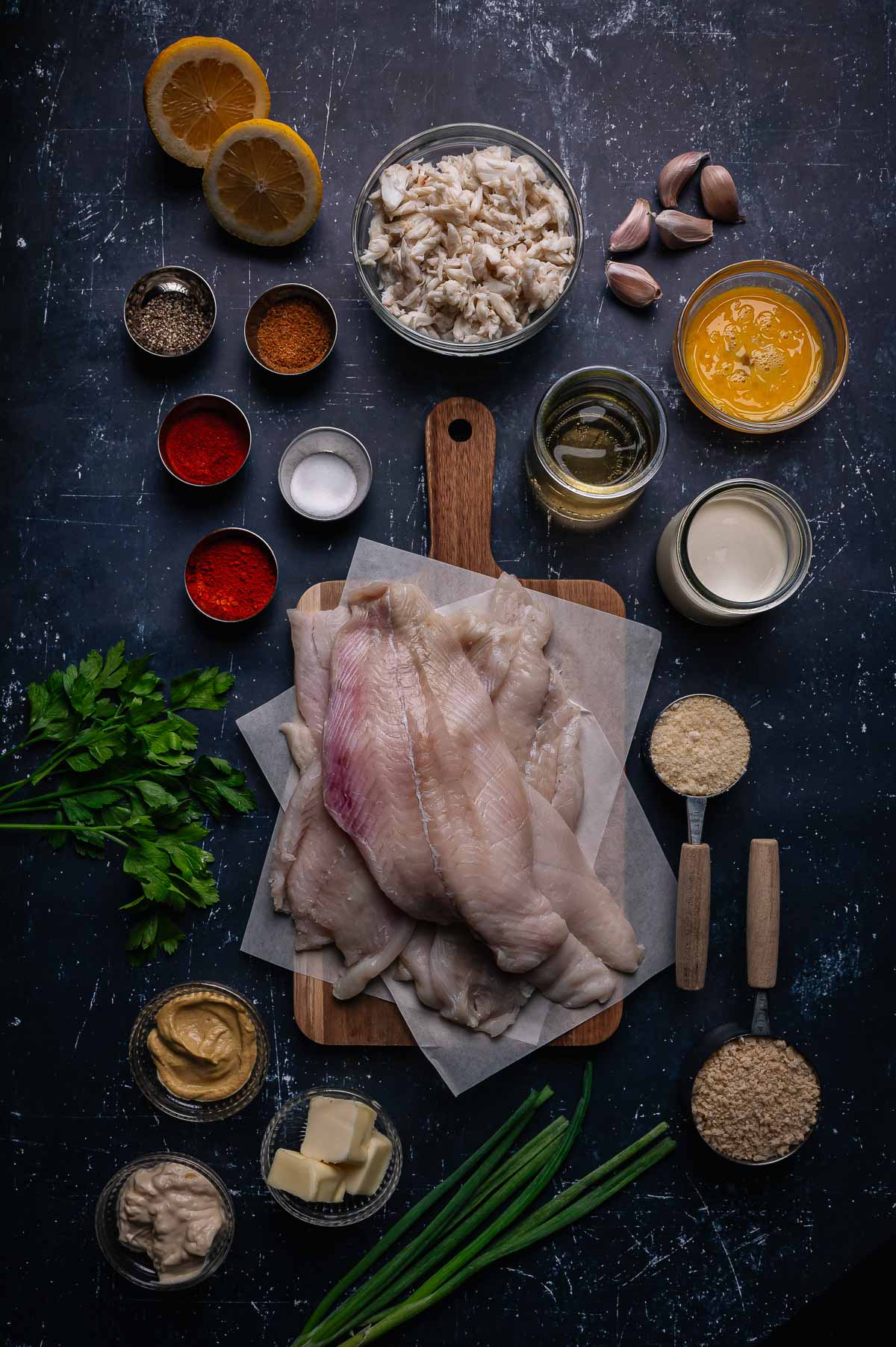 Overhead view of raw flounder fillets on a cutting board, surrounded by various ingredients including spices, crab meat, eggs, butter, lemon, parsley, and bowls of seasonings.